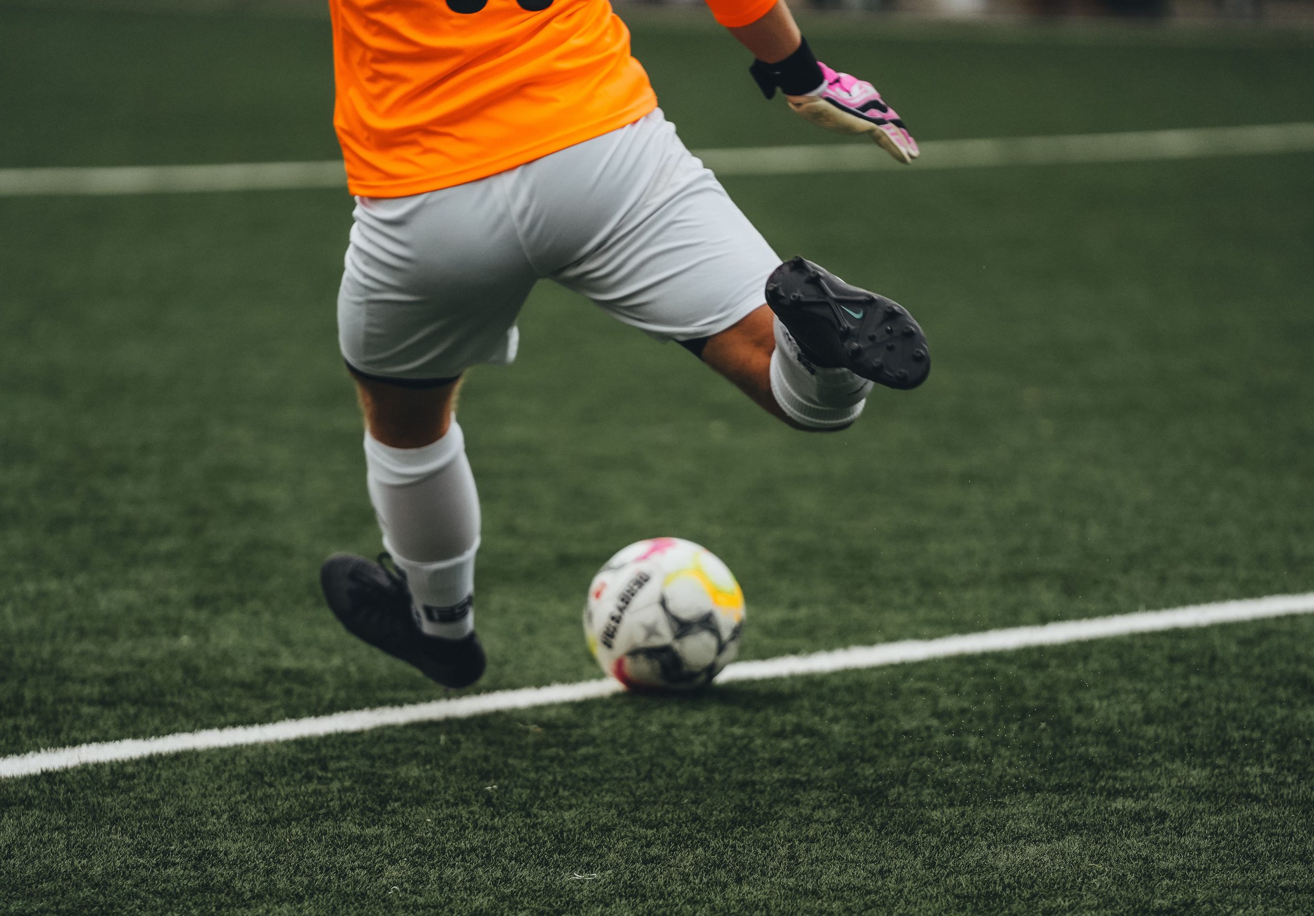 Footballer in an orange shirt striking a ball on artificial turf