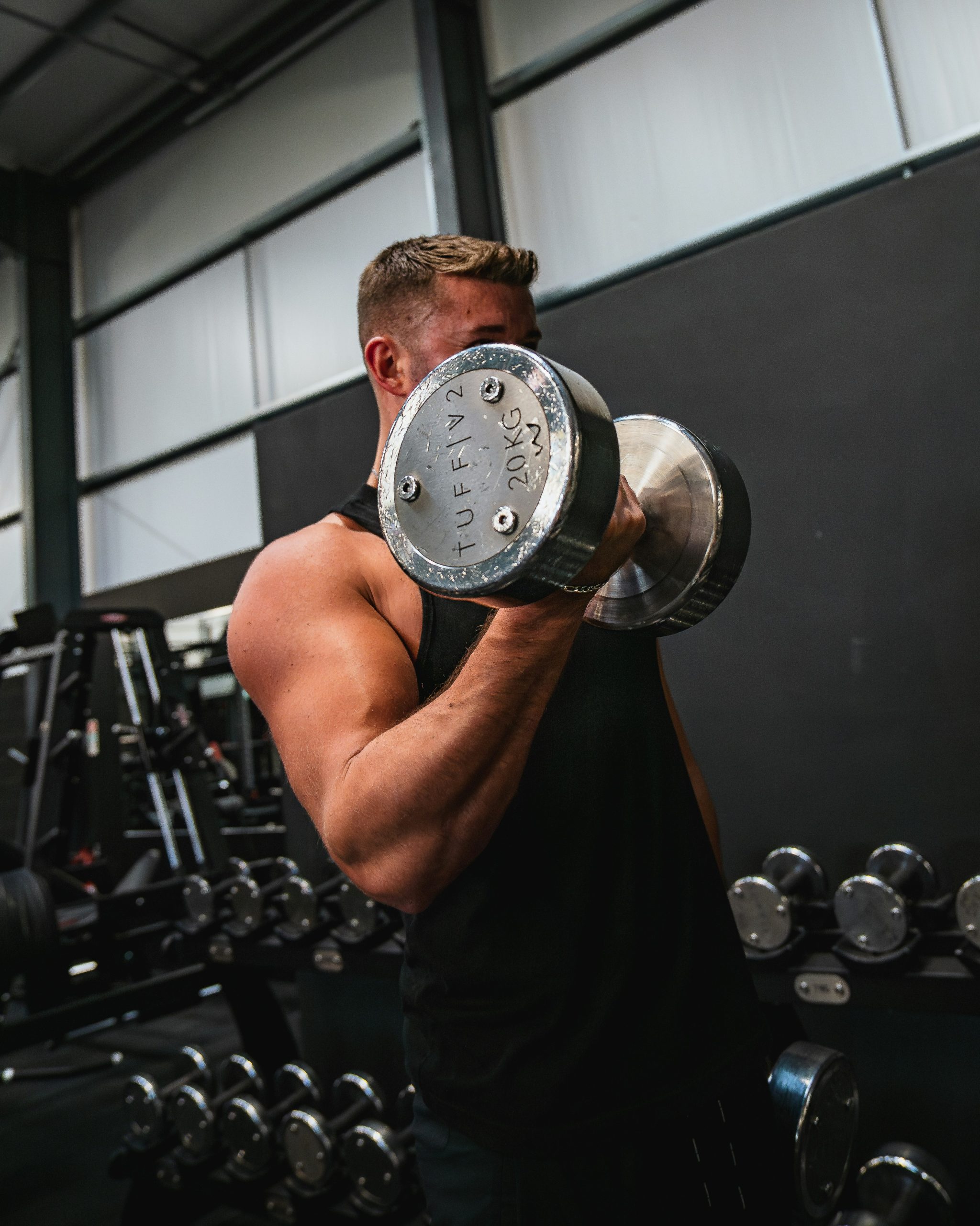 Man in a black tank top curling a heavy dumbbell, with weight racks behind him