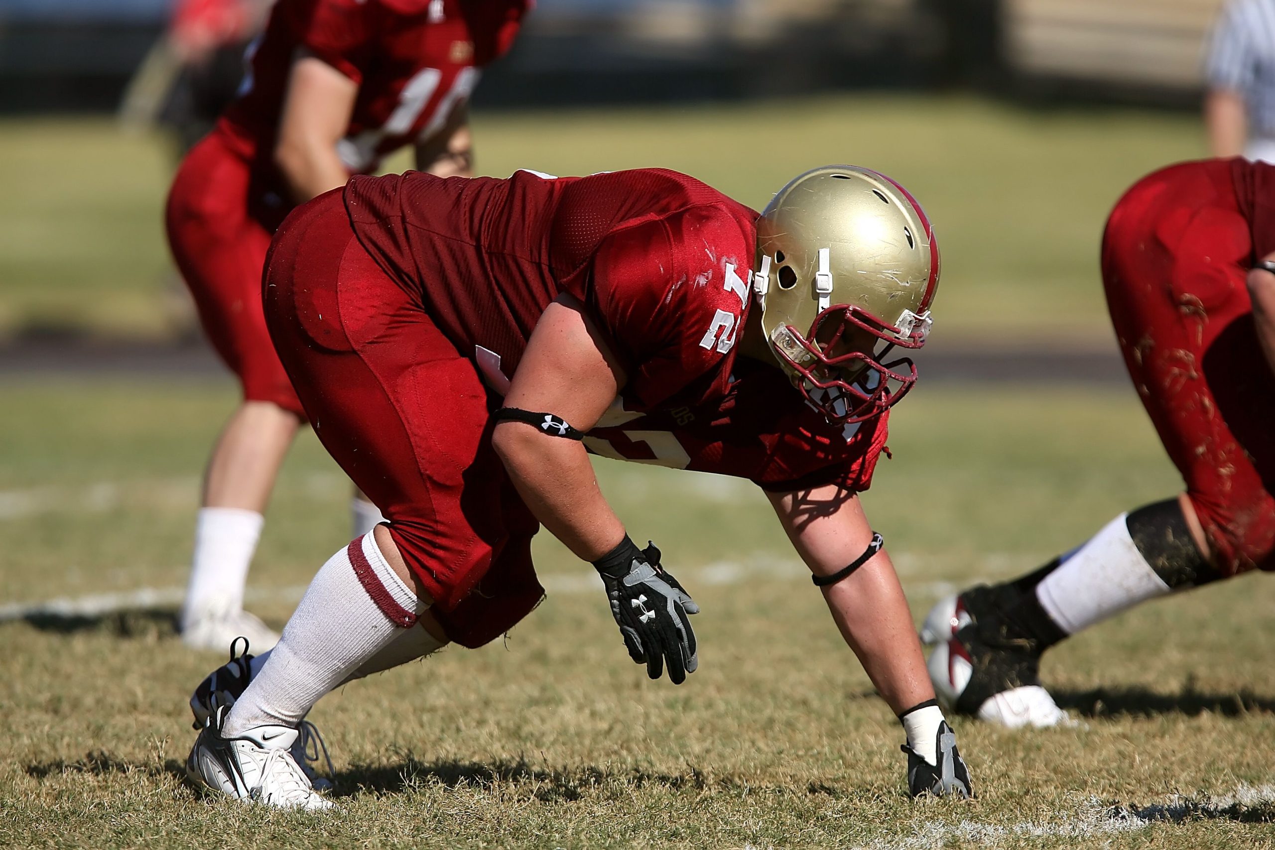 American football player in a red kit and gold helmet, crouched in a three-point stance on the field